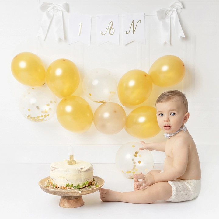 S&eacute;ance photo anniversaire b&eacute;b&eacute; &agrave; Toulouse, b&eacute;b&eacute; d&rsquo;un an assis devant un g&acirc;teau smash cake, d&eacute;cor de ballons dor&eacute;s et blancs en studio