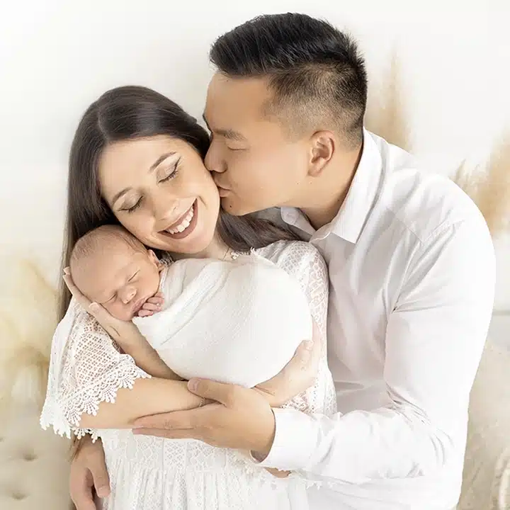 Un couple avec on b&eacute;b&eacute; et le papa qui embrasse la maman lors d'une s&eacute;ance photo naissance au studio de Tiana Photographe Toulouse